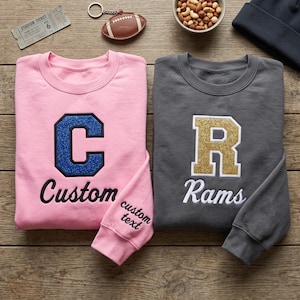 Peut inclure: Un sweat-shirt rose clair avec une lettre C à paillettes bleues et le mot Custom, et un sweat-shirt gris foncé avec une lettre R à paillettes dorées et le mot Rams. La manche rose porte le texte brodé custom text.