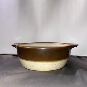 May include: A ceramic serving bowl with a two-tone design. The top half is a rich brown, while the bottom half is a speckled cream color. It has two small handles on either side. This bowl is suitable for serving soups, stews, or side dishes.