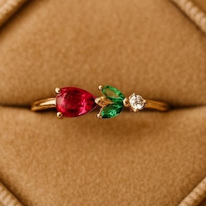 May include: A gold ring with a pear-shaped red gemstone, two green leaf-shaped gems, and a small white gem. The ring is set in a gold band and is displayed in a brown velvet ring box. The ring's design resembles a flower.