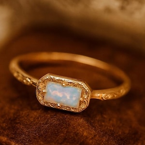 Puede incluir: Un anillo de oro con una piedra de ópalo. El ópalo rectangular está engastado en un marco dorado decorativo. La banda presenta un diseño ornamentado. El anillo se muestra sobre una superficie marrón.