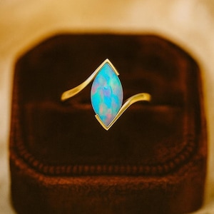 Può includere: Un anello d'oro con un'opale a forma di marquise con sfumature blu e viola. L'anello è incastonato in una fascia d'oro e presentato in un portagioie di velluto rosso scuro. Il design dell'anello è elegante e moderno.