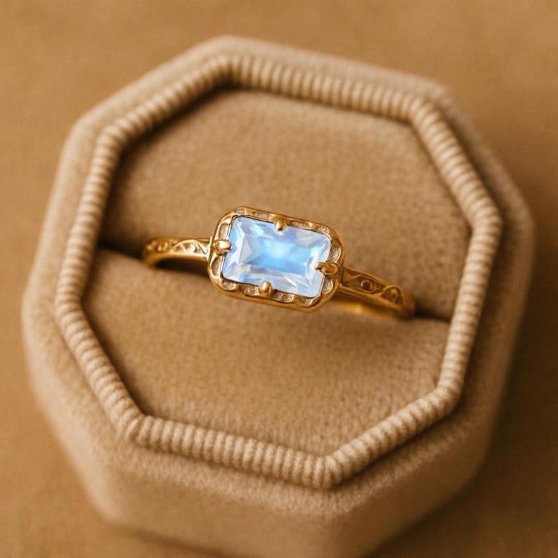 May include: A gold ring with a rectangular, light blue gemstone. The ring has an ornate, filigree design on the band and around the gemstone setting. The ring is displayed in a tan ring box.