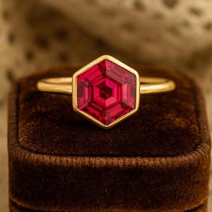 Pode incluir: Um anel de ouro com uma pedra preciosa vermelha rubi hexagonal e facetada. O anel é cravejado em uma banda de ouro e é exibido em uma caixa de anel de veludo marrom. A pedra preciosa tem um design geométrico.