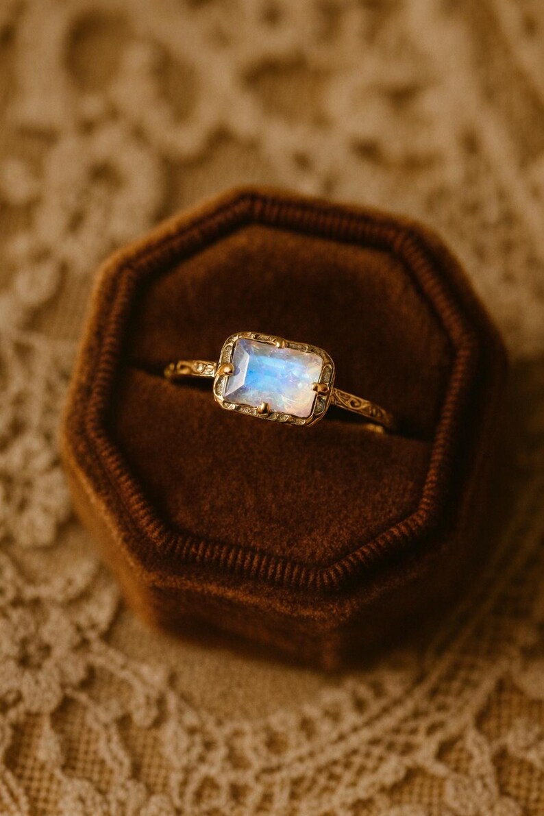 May include: A gold ring with a rectangular, iridescent gemstone, set in a decorative bezel. The ring is displayed in a brown velvet ring box, showcasing the intricate details of the jewelry.