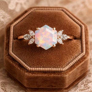 Könnte beinhalten: Ein roségoldener Ring mit einem sechseckigen Opal-Edelstein als Mittelpunkt. Der Opal hat einen schillernden Glanz. Flankiert wird der Opal von Gruppen kleiner, klarer Edelsteine. Der Ring liegt in einer braunen Samt-Schmuckschatulle.