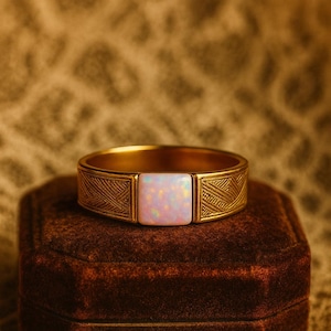 Könnte beinhalten: Ein goldfarbener Ring mit einem rechteckigen, opalisierenden Stein in der Mitte. Das Band weist ein geätztes geometrisches Muster auf. Der Ring liegt auf einer dunkelbraunen Samt-Schmuckschatulle vor einem strukturierten Hintergrund.