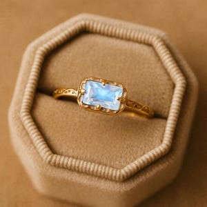 May include: A gold ring with a rectangular, light blue gemstone. The ring has an ornate, filigree design on the band and around the gemstone setting. The ring is displayed in a tan, octagonal ring box.