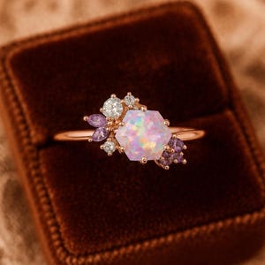 Könnte beinhalten: Roségoldener Ring mit einem großen, schillernden, sechseckigen Opal-Mittelstein. Der Opal ist von kleinen weißen und lila Edelsteinen umgeben, die ein Cluster-Design bilden. Der Ring befindet sich in einer Samt-Schmuckschatulle.