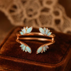 May include: A rose gold ring featuring a double band design. The ring is adorned with clusters of oval-shaped, opalescent gemstones, arranged in a symmetrical pattern. The gemstones have a soft, light blue hue, set against the warm tones of the metal.