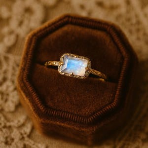 May include: A gold ring with a rectangular, iridescent gemstone, set in a decorative bezel. The ring is displayed in a brown velvet ring box, showcasing the intricate details of the jewelry.
