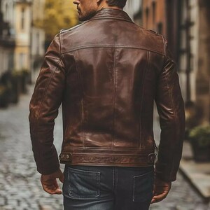 May include: A brown leather jacket with a classic design. The jacket features a notched collar, a zipper closure, and multiple pockets. The leather has a distressed look, giving it a vintage feel. The jacket is worn over a pair of blue jeans.