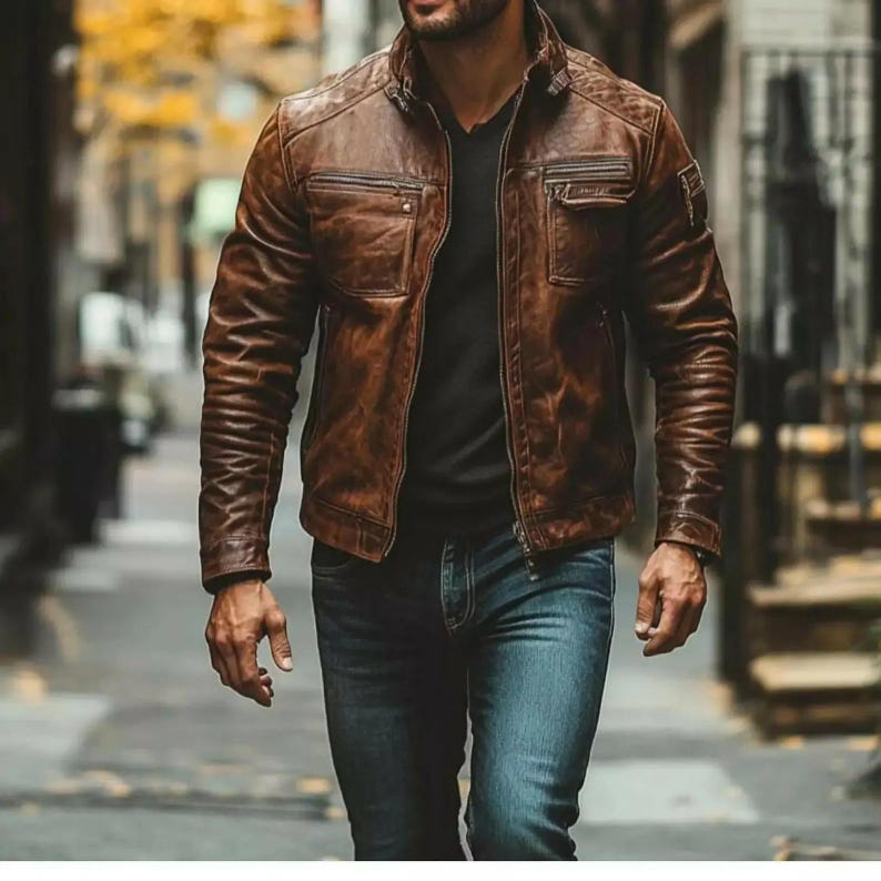 May include: A brown leather jacket with a classic design, featuring a stand-up collar, multiple pockets, and a zip-up front. The jacket is worn with a black shirt and blue jeans. The image is taken outdoors.