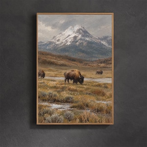May include: A framed landscape painting depicting three brown bison grazing in a grassy field with a small stream. In the background, a large, snow-capped mountain rises under a cloudy, overcast sky, rendered in muted earth tones and white.