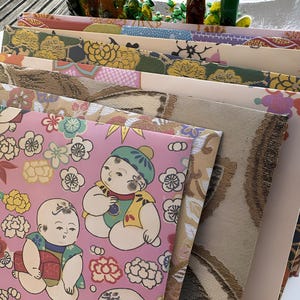 May include: A collection of patterned papers with diverse designs. The papers display floral, geometric, and illustrated patterns in shades of pink, gold, green, and beige. One paper has a pink background with illustrations of babies and flowers.