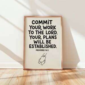 Op de afbeelding: Een rechthoekige wandprint in een lichthouten lijst met de zwarte tekst COMMIT YOUR WORK TO THE LORD. YOUR PLANS WILL BE ESTABLISHED. PROVERBS 16:3. Onder de tekst staat een zwarte lijntekening van twee handen die papier overhandigen.