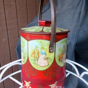 May include: Vintage red metal tin with a hinged lid and a brown handle. The tin features a decorative design with images of people in a garden setting. The tin is adorned with floral accents and a green and red color scheme.