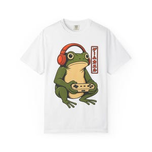 Op de afbeelding: Een wit t-shirt met een grafische illustratie van een groene kikker die een oranje koptelefoon draagt en een beige videospelcontroller vasthoudt. Rechts van de kikker staat een verticaal rood rechthoekig kader met Japanse tekst.
