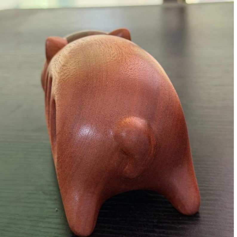 K&ouml;nnte beinhalten: Handgefertigte Holzskulptur eines stilisierten Tieres, m&ouml;glicherweise ein B&auml;r oder ein Schwein, von hinten betrachtet. Die Skulptur hat eine warme, r&ouml;tlich-braune Farbe mit glatten, polierten Oberfl&auml;chen und abgerundeten Merkmalen. Ohren und Beine sind sichtbar.