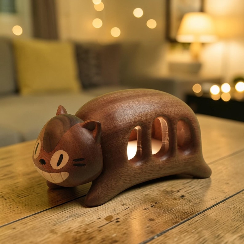 K&ouml;nnte beinhalten: Nachtlicht in Form einer Katze aus dunklem Holz. Die Katze hat ein l&auml;chelndes Gesicht und drei ovale Aussparungen. Sie hat spitze Ohren und einen Schwanz. Auf einem Holztisch platziert, mit verschwommenem Hintergrund und sanfter Beleuchtung.