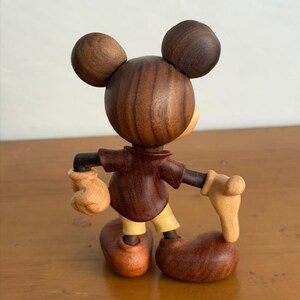 May include: Wooden Mickey Mouse figurine with a dark brown body, ears, and shoes. The figure has tan gloves, a yellow face, and yellow pants. The figurine is posed with its arms out and is standing on a wooden surface.