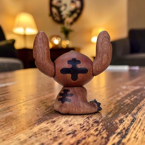 May include: A wooden figurine of a cartoon character with large ears and a cross-shaped design on its body. The figurine is brown with black accents and is posed on a wooden surface. The character's design is reminiscent of a popular animated film.