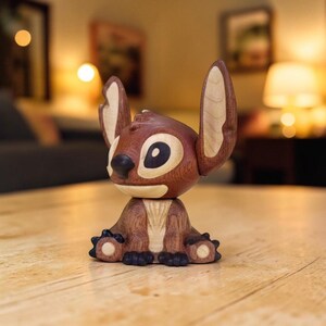 May include: A wooden figurine of the character Stitch from the animated film Lilo & Stitch. The figurine is primarily brown with lighter wood accents on the ears and face. It is sitting on a wooden surface.