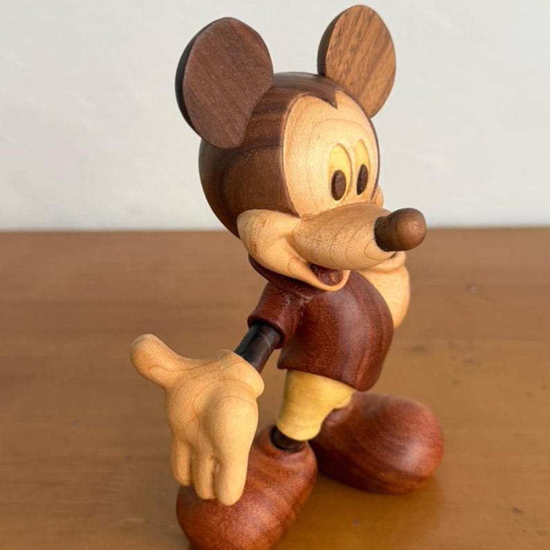 May include: A wooden Mickey Mouse figurine. The figure is crafted from various shades of wood, with a dark brown head and ears, a lighter brown face and hands, and a reddish-brown body. The figure is posed with one hand extended.