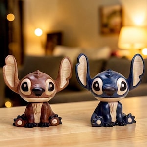 May include: Two wooden Stitch figurines. One is brown with light brown ears, the other is blue with light blue ears. Both have large black eyes and are sitting down. The figurines are on a wooden surface.