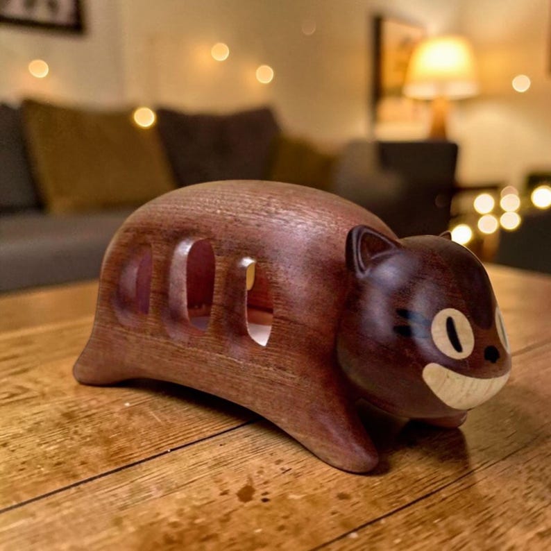 K&ouml;nnte beinhalten: Eine handgeschnitzte Holzkatzenfigur in Braun. Die Katze hat ein l&auml;chelndes Gesicht mit schwarzen Details und drei rechteckigen Aussparungen an der Seite. Sie steht auf einer Holzoberfl&auml;che.