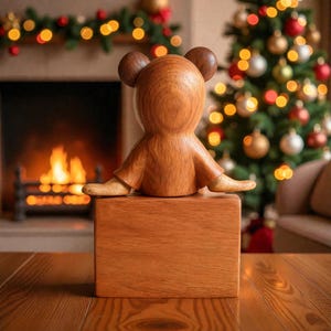 Pu&ograve; includere: Una statuina di legno a forma di orso con orecchie arrotondate e braccia tese, seduta su un blocco di legno. La statuina &egrave; di colore marrone caldo. Lo sfondo comprende un albero di Natale con ornamenti e un camino.