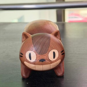 K&ouml;nnte beinhalten: Eine Holzkatzenfigur mit einem l&auml;chelnden Gesicht, gro&szlig;en ovalen Augen und kleinen Ohren. Die Katze ist braun mit einem dunkleren braunen Streifen auf dem Kopf und helleren Holzakzenten im Gesicht. Die Figur ist ein dekoratives Objekt.