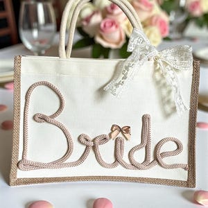 May include: A cream-colored tote bag with the word "Bride" in pink rope lettering. The bag has rope handles and a decorative lace bow. The edges are trimmed with a burlap-like material. Pink rose petals are scattered around the bag.