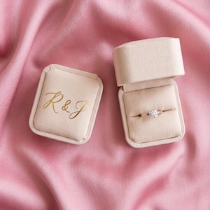 May include: Two beige velvet ring boxes on a pink satin background. One box is open, displaying a diamond ring. The other box is closed, with the gold letters "R&J" on the lid. The ring has a silver band and a large, square-cut diamond.