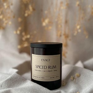 Peut inclure: Un pot de bougie noir avec un couvercle, étiqueté "EXACT SPICED RUM". L'étiquette indique les ingrédients : fève tonka, rhum, amande et miel. La bougie est coulée à la main à Lisbonne et fabriquée avec de la cire de soja. Le pot est posé sur un tissu de couleur neutre.