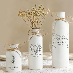 Puede incluir: Tres jarrones de cerámica blanca de diferentes tamaños, cada uno adornado con un gráfico negro y atado con hilo. Los jarrones presentan diseños botánicos y geométricos, con nombres y fechas. Un jarrón contiene flores secas.