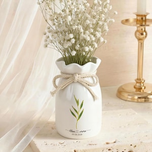 May include: A white ceramic vase with a ruffled top and a beige rope tie, holding a bouquet of white flowers. The vase has a green leaf design and the text "ethan & emma 05 06 2026".
