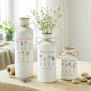 May include: Three white ceramic vases with gold rims and twine bows. Each vase features floral illustrations and the text "Nana's Garden" and "Love Grows Here". One vase holds flowers.