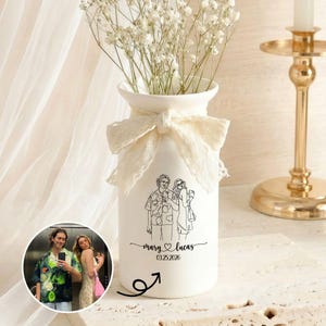 Puede incluir: Un jarrón de cerámica blanca con un lazo de encaje y un dibujo lineal de dos personas. El jarrón está lleno de flores blancas. El texto "mary & lucas 03.25.2026" está debajo del dibujo. Una foto de la pareja está en un círculo.