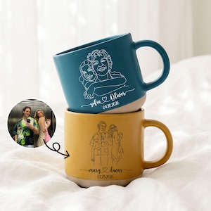 May include: Two ceramic mugs stacked on top of each other. The top mug is teal with a white line drawing of three people and the names "sofia & oliver" and the date "04.20.2026". The bottom mug is yellow with a white line drawing of three people and the names "mary & lucas" and the date "03.25.2026".