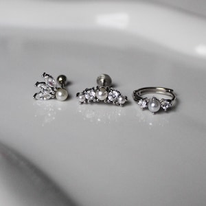 Könnte beinhalten: Ein Set aus drei silberfarbenen Schmuckstücken mit Perlen und klaren Kristallen. Das Set beinhaltet einen Ohrstecker mit Perle und Kristallakzenten, einen gebogenen Barbell-Ohrring und einen Ring mit einer tropfenförmigen Perle.