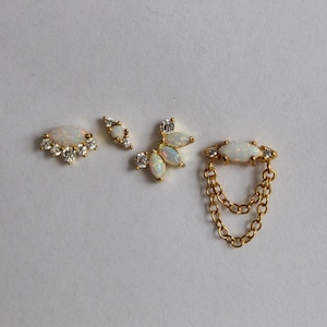May include: Four pieces of gold-toned jewelry with opal and clear crystal accents. The pieces include a stud with a fan-shaped design, a small stud, a cluster stud, and a double-chain earring.
