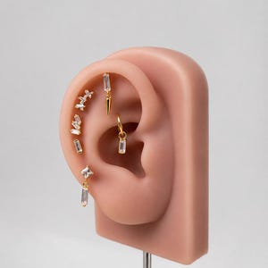 Könnte beinhalten: Ein Set goldfarbener Ohrringe auf einem synthetischen Ohrmodell. Die Kollektion umfasst mehrere kleine Stecker mit klaren Baguette-Steinen, einen hängenden Dorn-Anhänger, einen kleinen Creolen-Ohrring und einen quadratischen Stecker mit Anhänger.