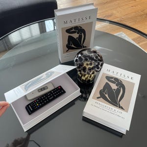 Op de afbeelding: Een stilleven met twee boeken met "MATISSE" op de omslag, een decoratieve glazen bol en een witte doos met twee afstandsbedieningen. De boeken tonen een zwart-beige illustratie.