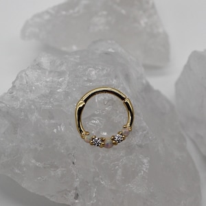 Op de afbeelding: Een goudkleurige scharnierende oorring met drie decoratieve elementen. De oorring heeft twee kleine, ronde, opalescente stenen en een centrale, heldere, sprankelende steen. De oorring is geplaatst tegen een achtergrond van witte, kristallijne formaties.