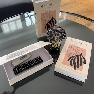 Könnte beinhalten: Ein dekoratives Arrangement mit zwei Büchern mit dem Titel "MATISSE THE CUT-OUTS" mit einem floralen Design, einer Glaskugel und einer Box mit zwei Fernbedienungen. Die Bücher haben einen beigefarbenen Einband mit einer rosa-schwarzen Illustration. Die Fernbedienungen sind schwarz und silber.