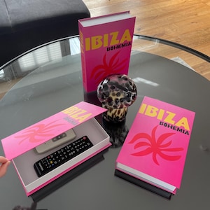Könnte beinhalten: Dekoratives Set mit zwei pinkfarbenen Boxen und einem passenden Buch, alle mit dem Text "IBIZA BOHEMIA" in Gelb. Eine Box ist geöffnet und zeigt eine Fernbedienung. Eine gemusterte Glaskugel befindet sich in der Nähe.