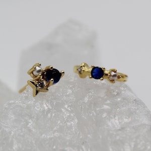Könnte beinhalten: Ein Paar goldfarbene Ohrringe und ein Ring. Die Ohrringe haben einen Stern, eine Perle, einen Diamanten und einen dunkelblauen Edelstein. Der Ring hat einen Diamanten, einen dunkelblauen Edelstein und eine Perle. Der Schmuck liegt auf einer weißen Oberfläche.