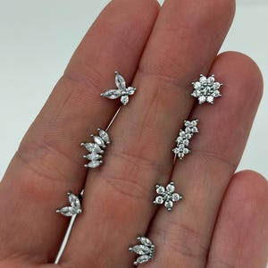 Può includere: Una collezione di orecchini argentati con pietre di zirconia cubica trasparenti e scintillanti. Gli orecchini presentano vari design, tra cui farfalle, fiori e foglie. Gli orecchini sono esposti su uno sfondo neutro.
