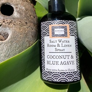 May include: A black bottle with a white label that reads "Salt Water Room & Linen Spray Coconut & Blue Agave Made with Aloha in Hawaii". The bottle is sitting on a green leafy surface.