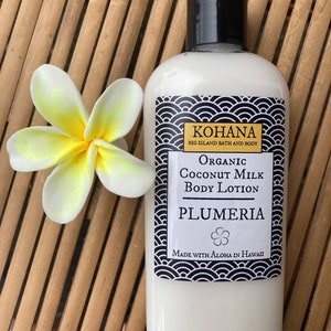 May include: A white bottle of Kohana Organic Coconut Milk Body Lotion with a black cap and a label featuring a black and white wave pattern. The label reads "Plumeria" and "Made with Aloha in Hawaii." A white and yellow plumeria flower rests beside the bottle.